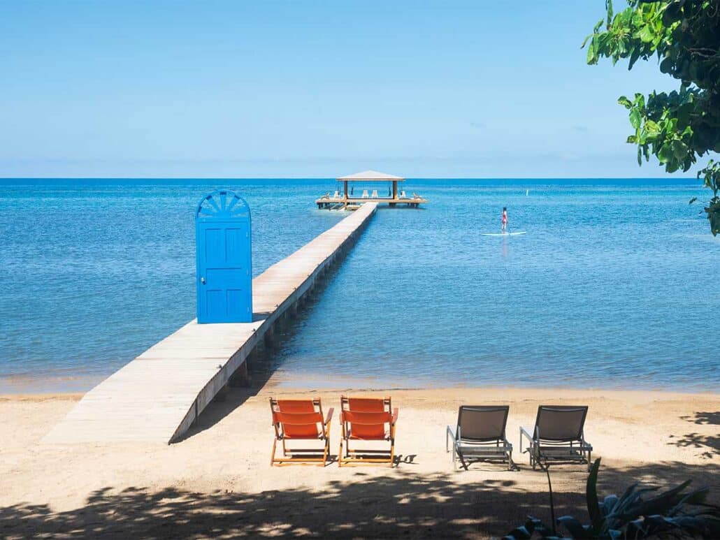 View of the private beach at Puerta Azul Roatan featuring orange lounge chairs, a paddleboarder in the calm sea, and the iconic blue door on the wooden pier leading to the ocean palapa.