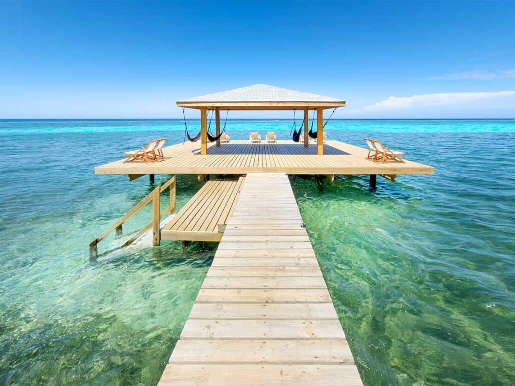 A wooden pier leading to a shaded palapa with hammocks and lounge chairs over the clear turquoise water at Puerta Azul Roatan