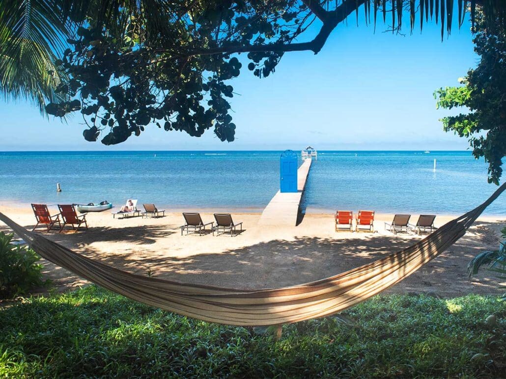 View from a hammock under tropical trees looking out toward the private beach, turquoise ocean, and the blue door pier at Puerta Azul Roatan.