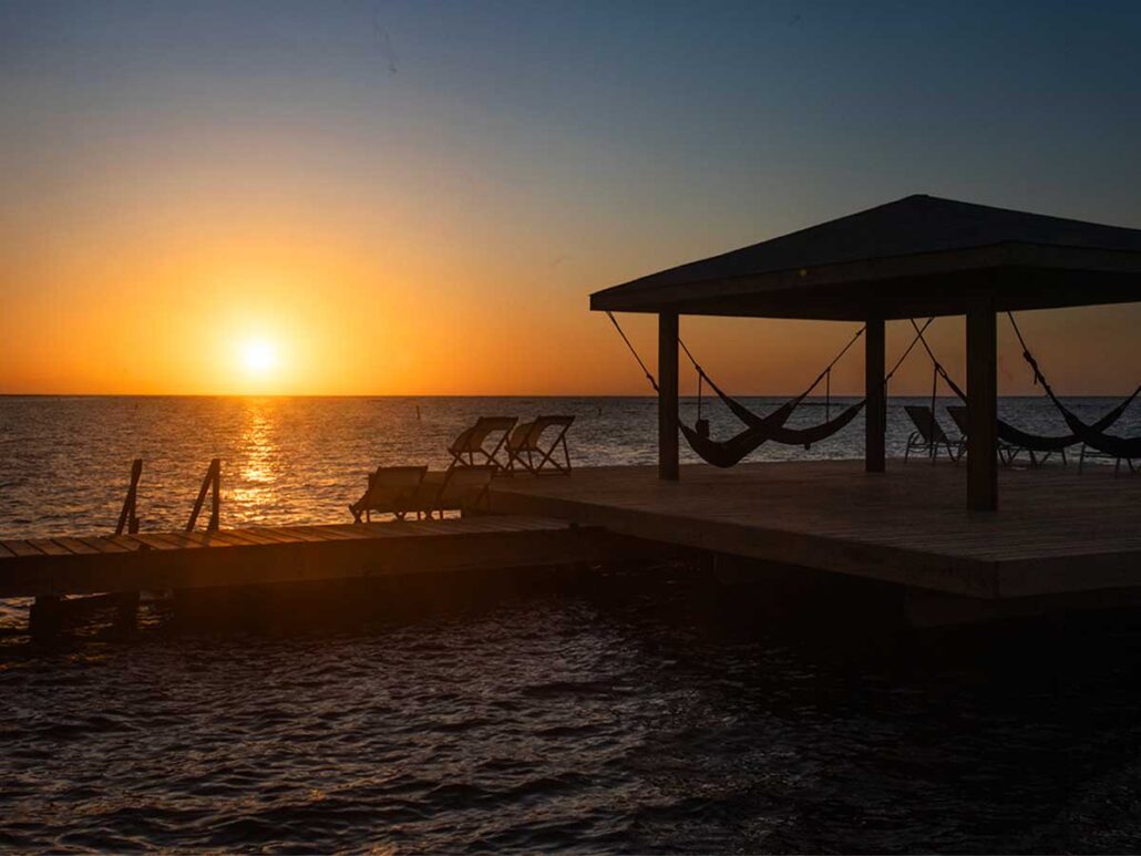 Sunset silhouette of the private over-water palapa and hammocks at Puerta Azul Roatan, with the sun reflecting on the Caribbean Sea.
