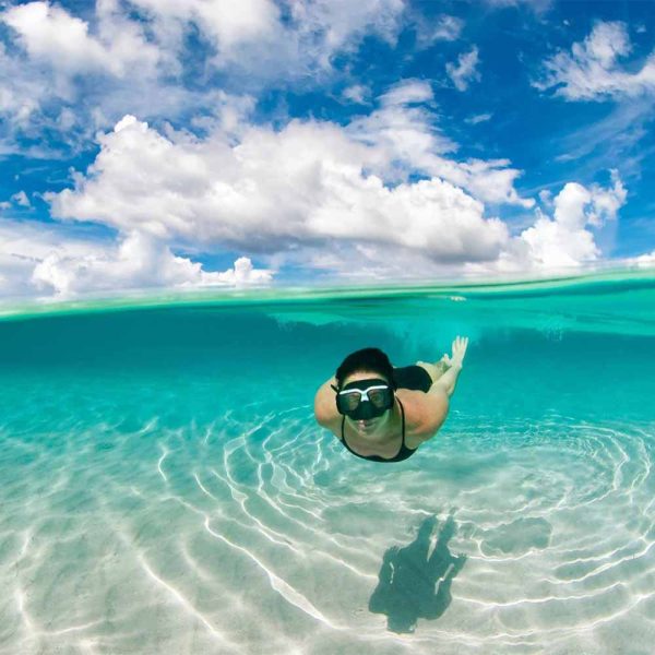 Split-level underwater and above-water view of a woman snorkeling in the crystal-clear turquoise waters of Pigeon Cay, Roatan, under a bright cloudy sky.