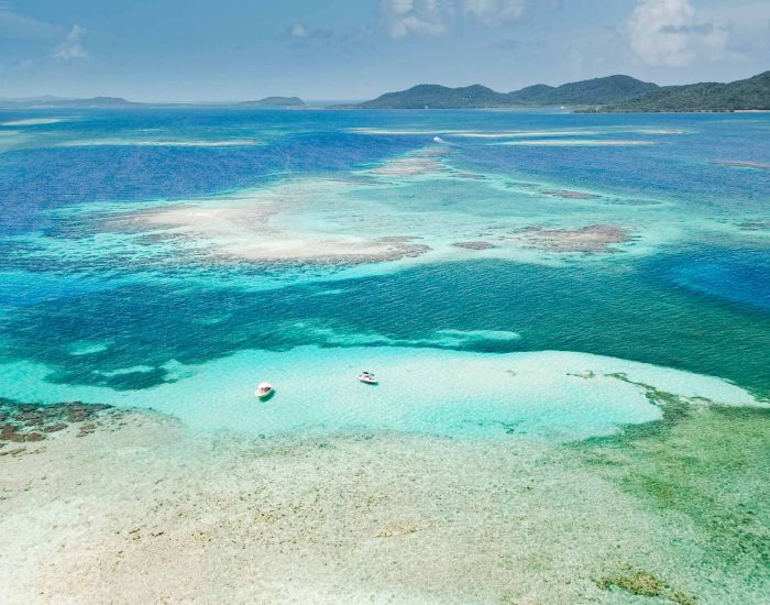 Aerial view of two small boats anchored in the crystal-clear turquoise waters of Pigeon Cay near Roatan, showcasing the vibrant coral reef and white sand bars.