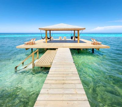 A wooden pier leading to a shaded palapa with hammocks and lounge chairs over the clear turquoise water at Puerta Azul Roatan