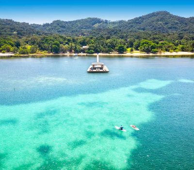 Wide aerial view of Puerta Azul Roatan hotel nestled in lush green hills with its private dock extending into the turquoise Caribbean Sea toward the reef.