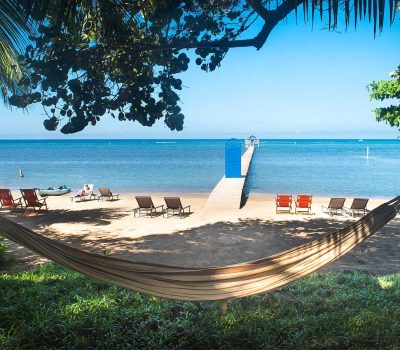 View from a hammock under tropical trees looking out toward the private beach, turquoise ocean, and the blue door pier at Puerta Azul Roatan.